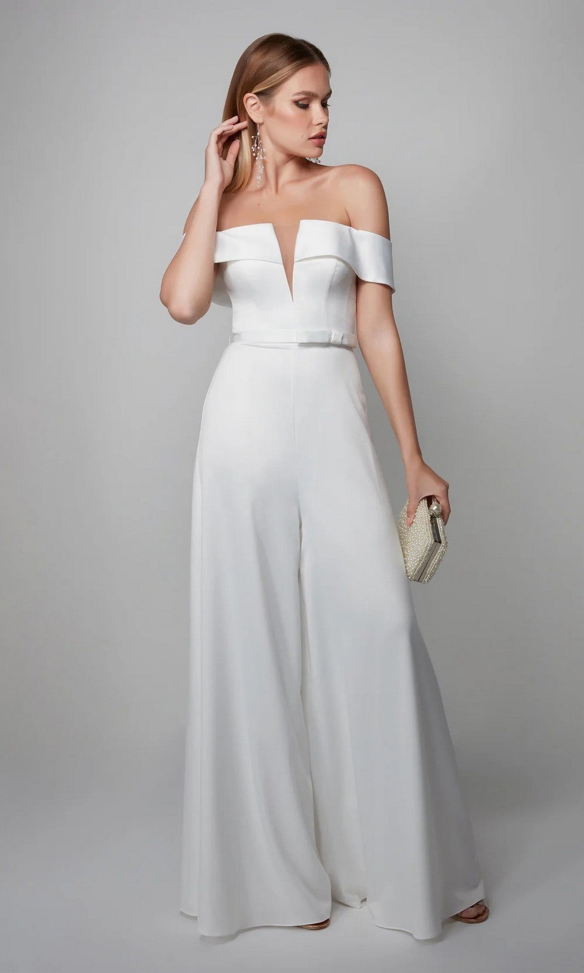Woman in the Alyce 70012 long formal jumpsuit featuring off-shoulder style, wide legs, and fitted waist, complemented by a textured clutch and long drop earrings, stands against a plain background.